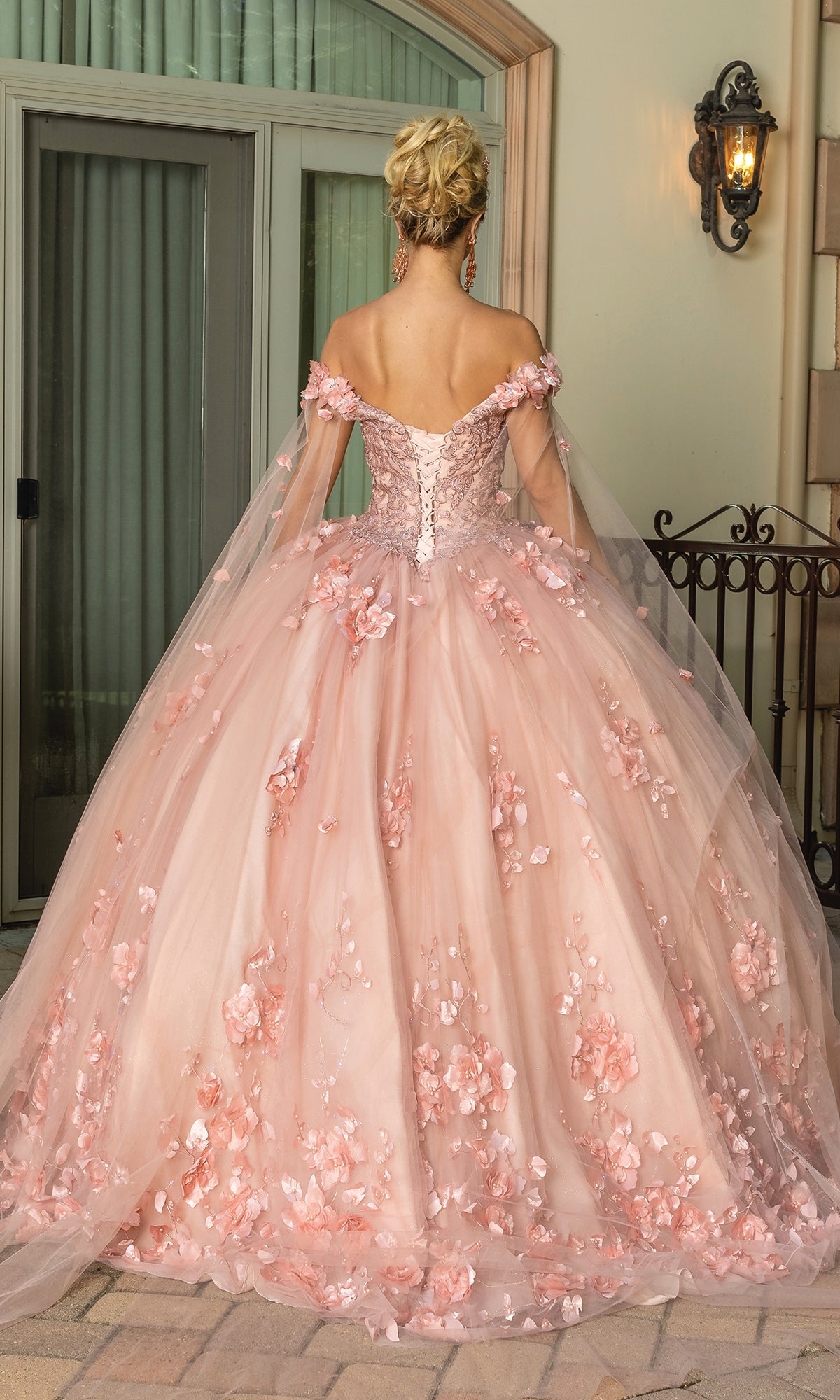 A woman with an elegant updo faces away in front of a doorway, wearing the Quinceanera Dress 1695 by Dancing Queen—a light pink, off-the-shoulder A-line gown adorned with floral appliqués.